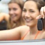 Girls are sitting in their car at the beach holding new keys out the window.
