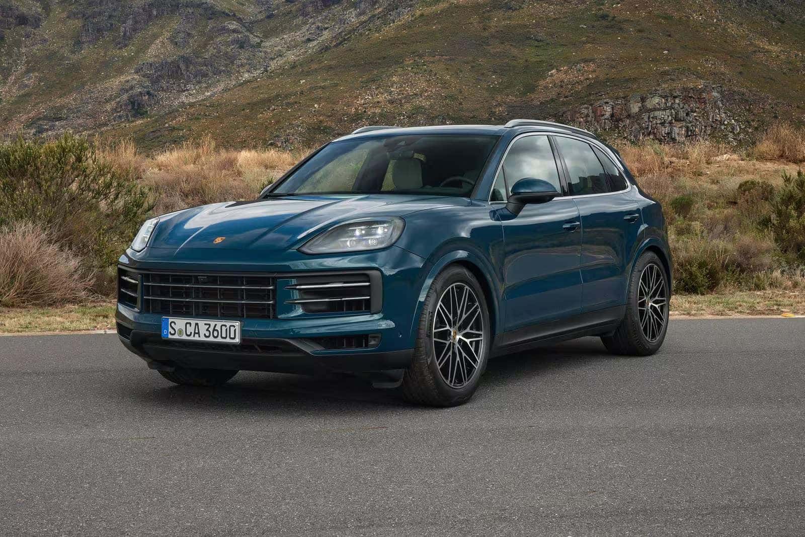 a blueish green 2025 porsche cayenne parked on a paved road with rolling hills in the background