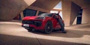 A man leans casually against a red Porsche Cayenne, parked in front of a modern architectural building with sharp, angular lines. The striking red color of the Porsche Cayenne contrasts with the neutral tones of the surrounding architecture, offering a visual comparison point for those evaluating the Porsche Macan vs Cayenne.