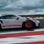 White porsche 911 gt3 rs driving on a racetrack with a large rear wing and red wheels under cloudy skies.