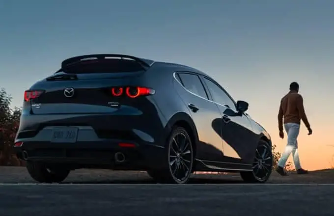 A black Mazda hatchback is parked on the side of the road, and a man is walking in front of it.